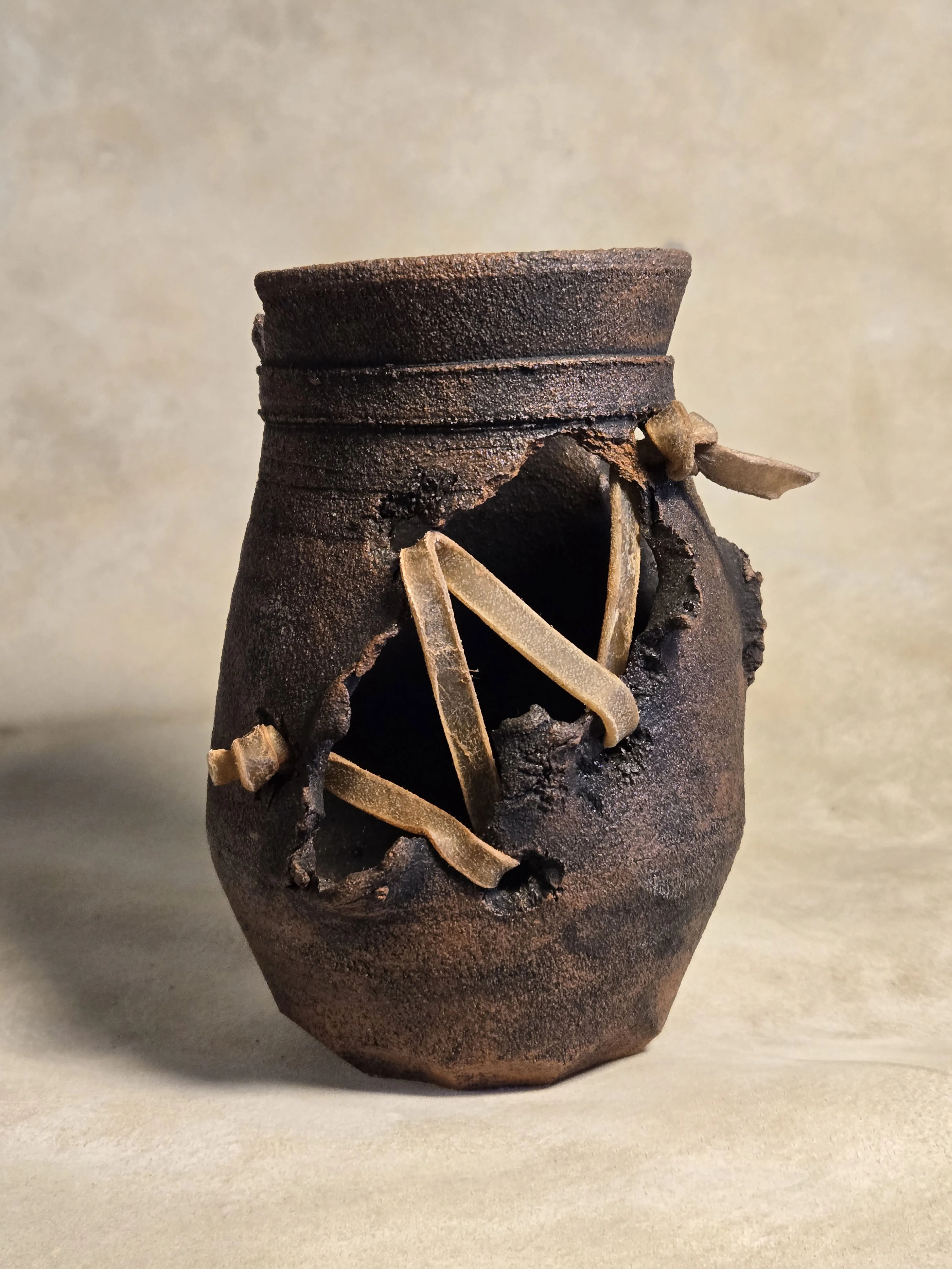 A wild-clay vessel torn open at the shoulder and laced back together with rawhide strips.