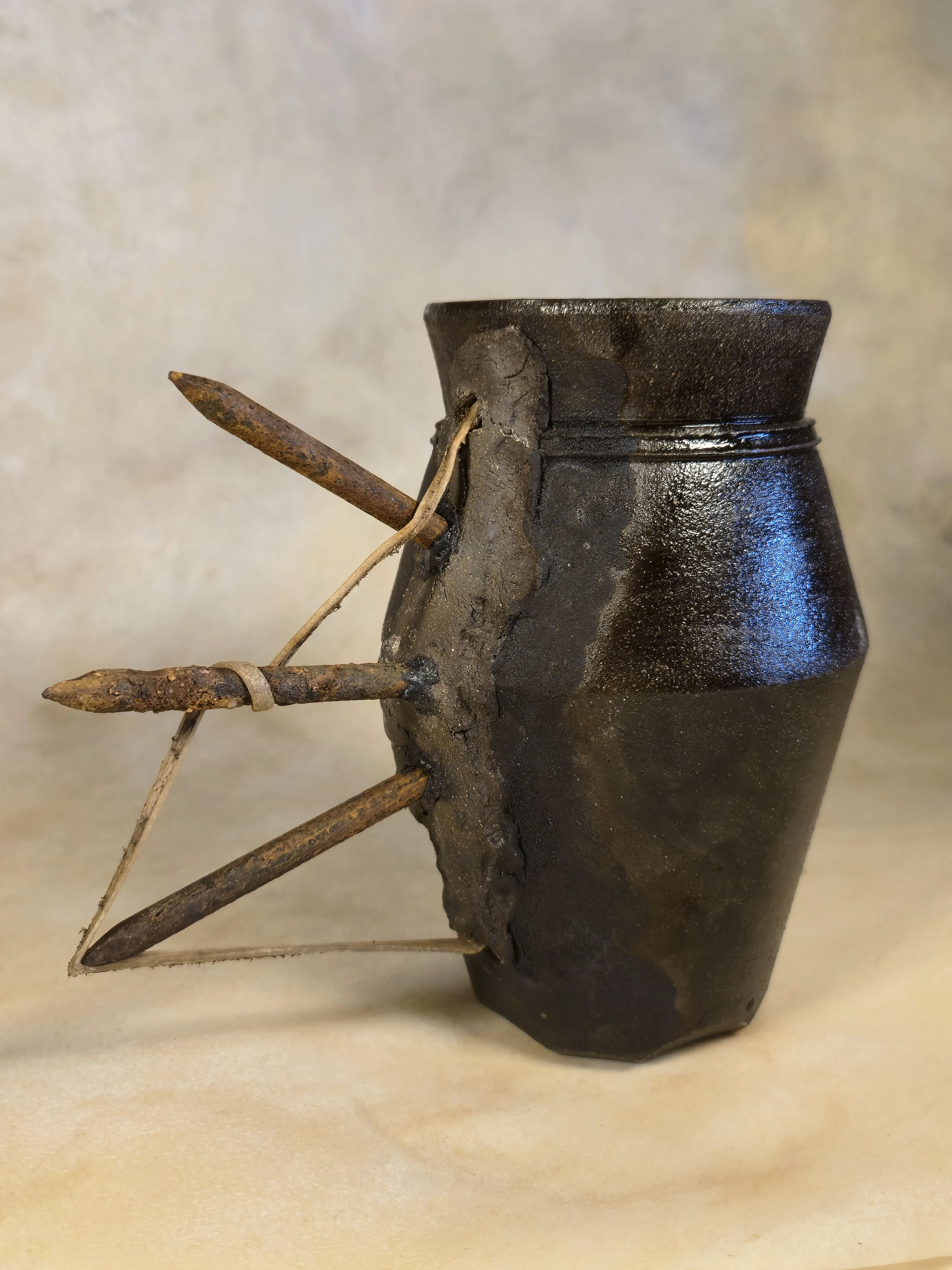 A glossy dark vessel with three rusted spikes driven through one side, bound with rawhide.