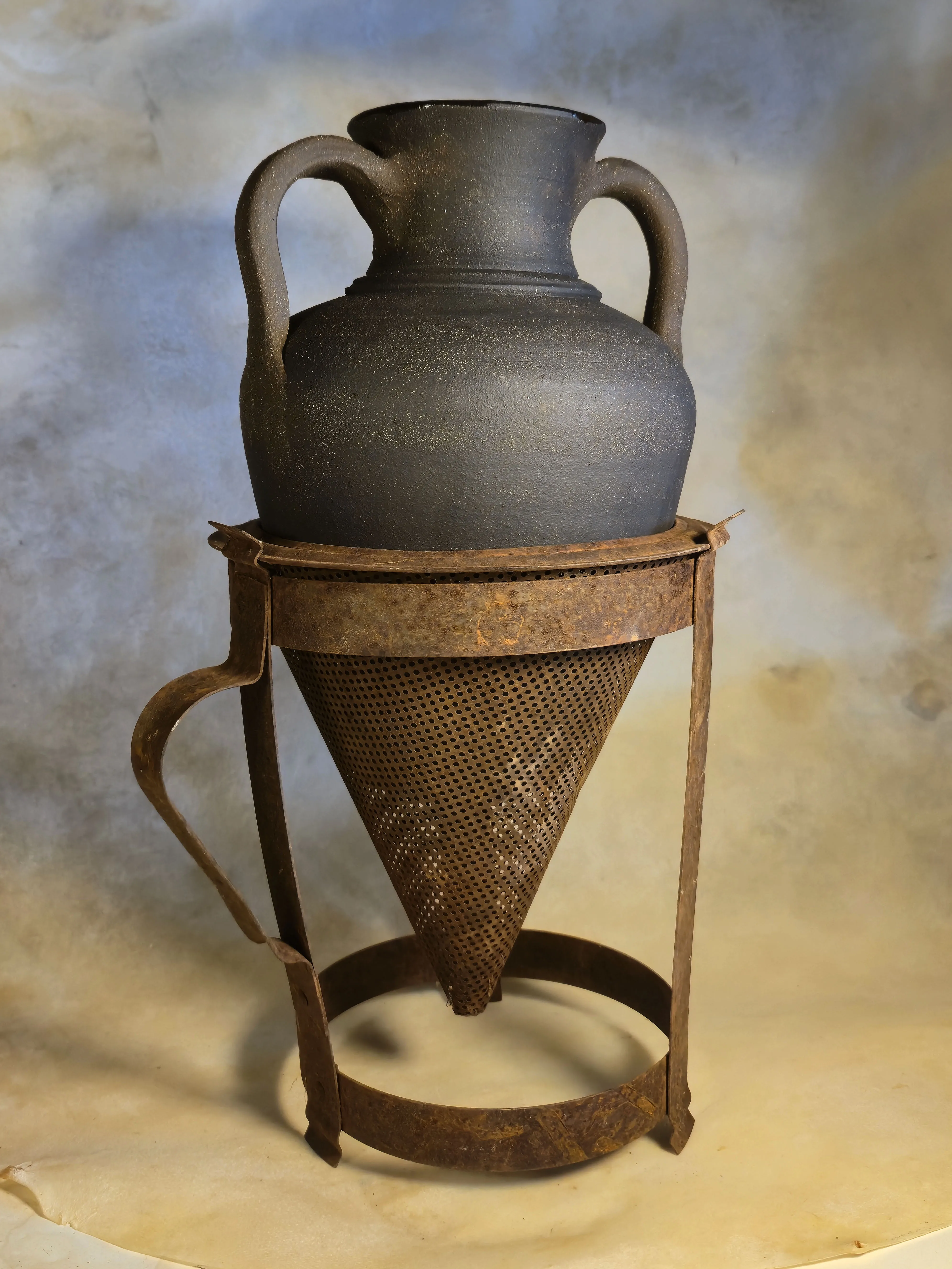 A dark two-handled amphora resting in a rusted 1930s cone-shaped fruit strainer on a steel stand.